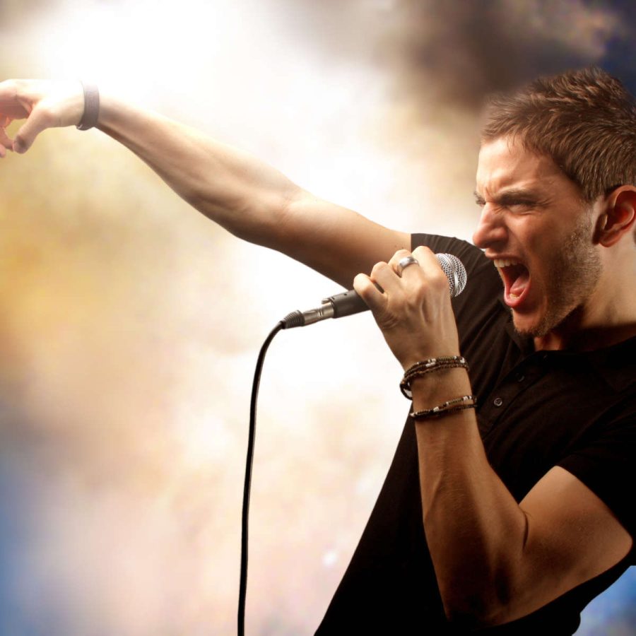 junger Sänger mit Dreitagebart auf der Bühne, der aufs Publikum zeigt vor rauchigem farbigem Hintergrund