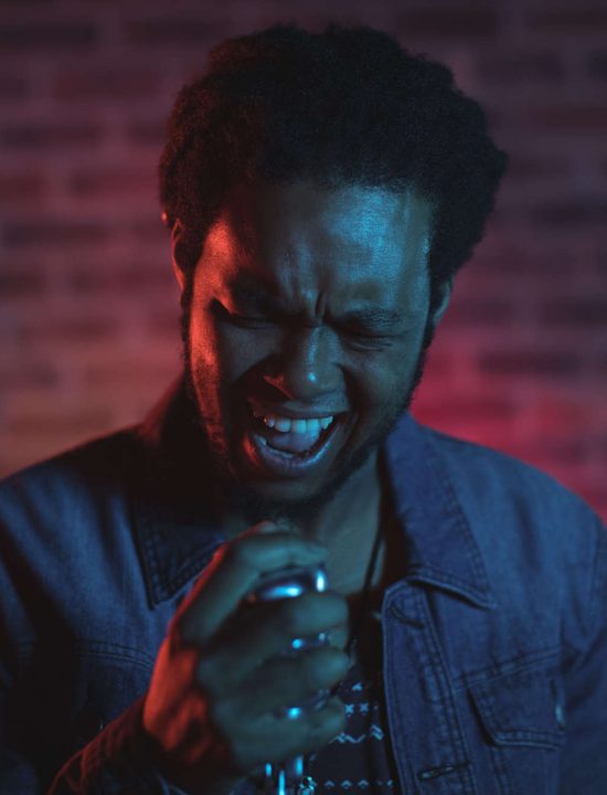 Sänger, der mit geschlossenen Augen den Kopf leicht geneigt ins Mikrofon singt vor einer rot angeleuchteten Backsteinwand