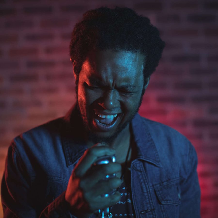 Sänger, der mit geschlossenen Augen den Kopf leicht geneigt ins Mikrofon singt vor einer rot angeleuchteten Backsteinwand